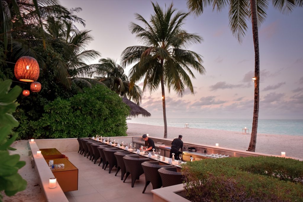 Außenbereich des Restaurants Koko Grill mit gedeckten Tischen aus dunklem Geflecht, Palmen und Meer im Hintergrund