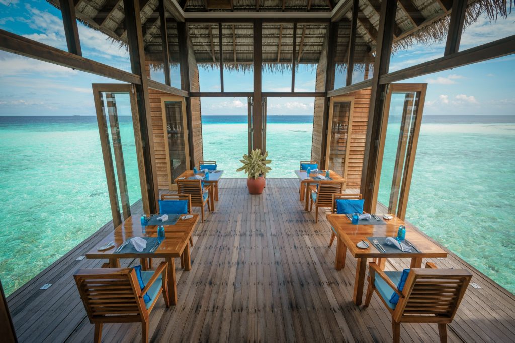 Offenes Restaurant Mandhoo mit Holztischen, blauen Kissen und Panoramablick auf türkisblaues Meer auf den