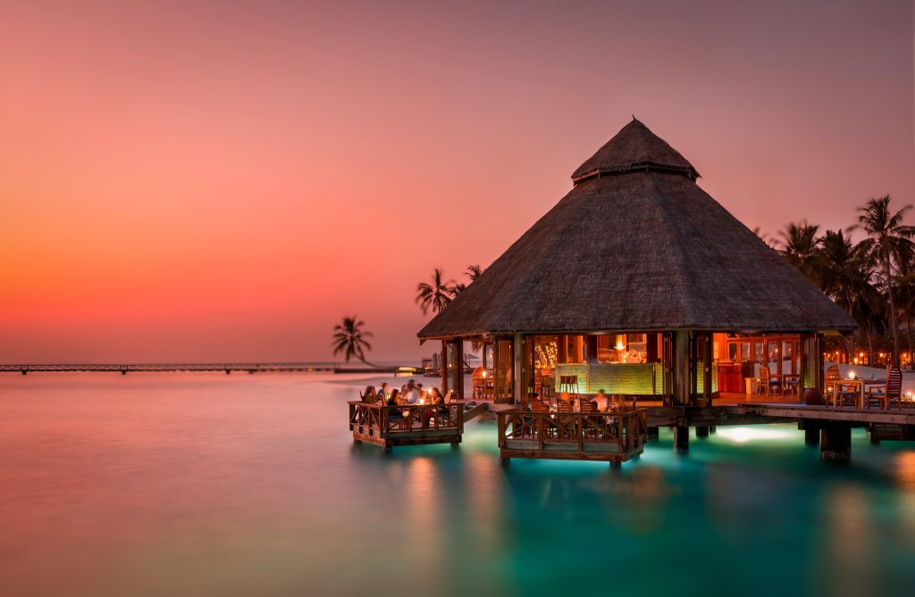 Palmenumsäumtes Wasserbungalow-Restaurant Sunset Grill bei Sonnenuntergang mit Holzdecks und Laternenlicht