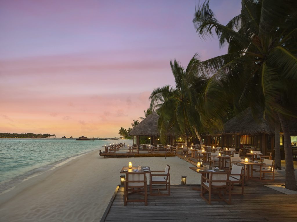 Strandterrasse des Restaurants Vilu mit Holztischen, weißen Polstern und Kerzen bei Sonnenuntergang auf den