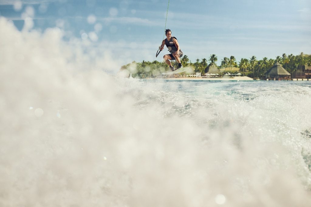 Wakeboarder springt über Wellen mit tropischen Palmen und Holzhütten auf Stelzen im Hintergrund auf den