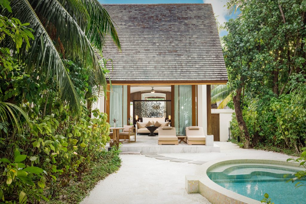 Deluxe Beach Villa mit Holzschindeldach, Glasfronten, zwei Liegestühlen und rundem Pool umgeben von Sand und Dschungel.