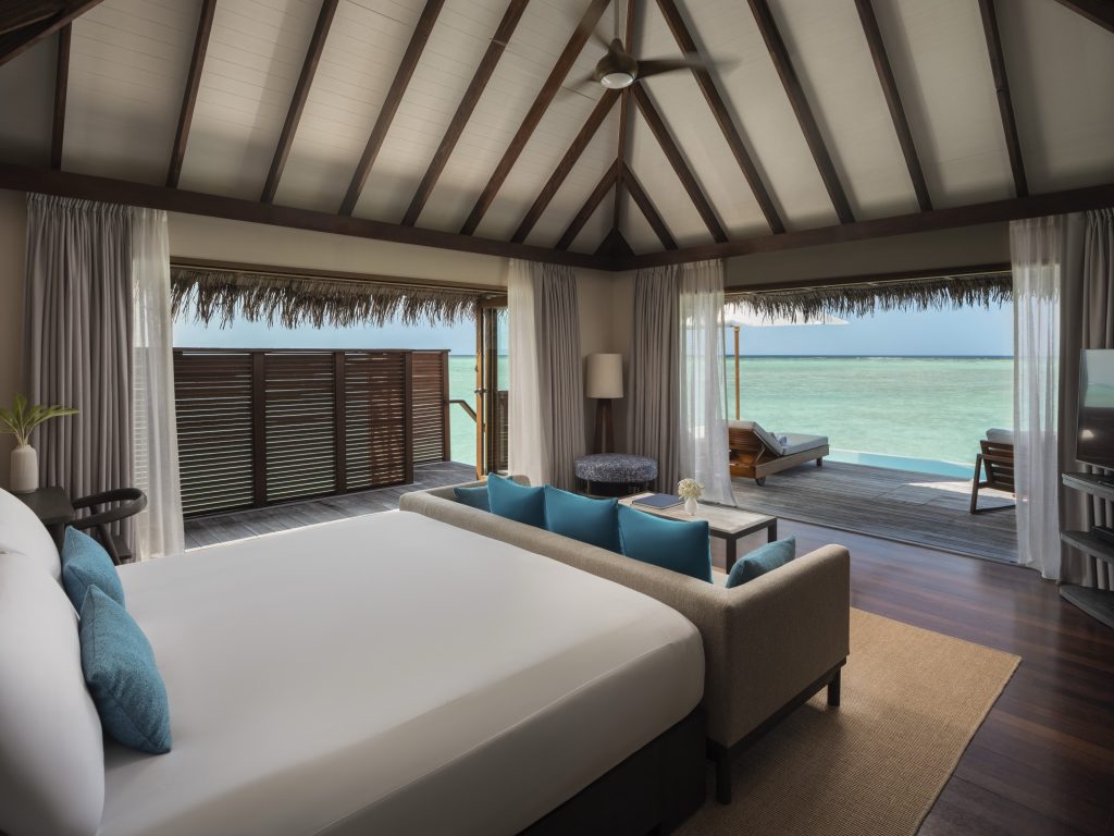 Zimmer der deluxe water villa mit großem Bett, türkisfarbenen Kissen, Holzfußboden und Meerblick auf den