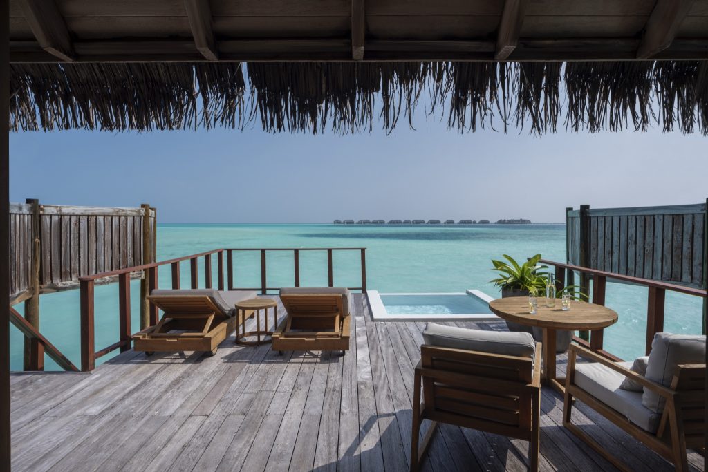 Terrasse eines Water Villa Zimmers mit Holzliegen, rundem Holztisch, Pool und Blick auf das türkisfarbene Meer