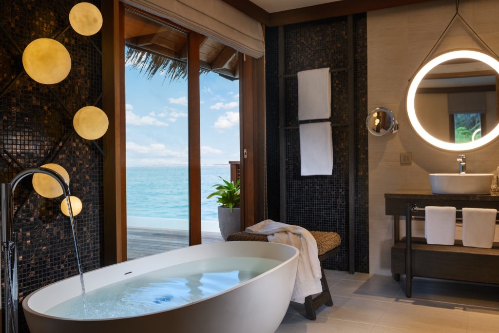 Badezimmer mit freistehender weißer Badewanne, dunklem Mosaik, rundem Spiegel und Meerblick im Conrad Maldives Rangali Island