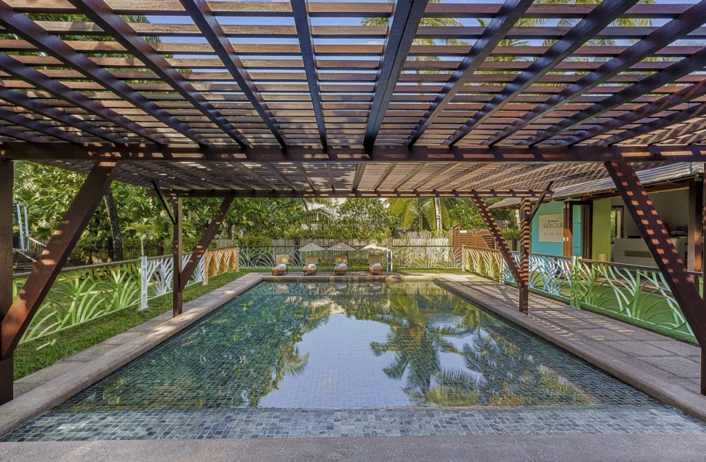 Rechteckiger Pool mit grünen Fliesen unter einer dunklen Holzpergola und Sonnenliegen im Hintergrund