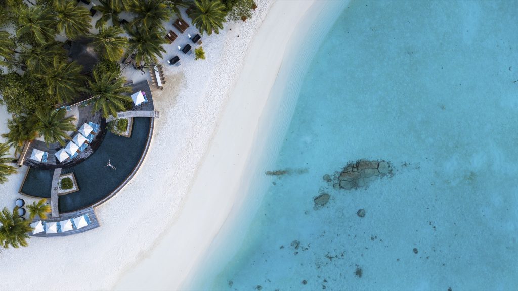 Luftaufnahme des Poolbereichs mit weißen Sonnenschirmen, Palmen und hellem Sandstrand am türkisfarbenen Meer