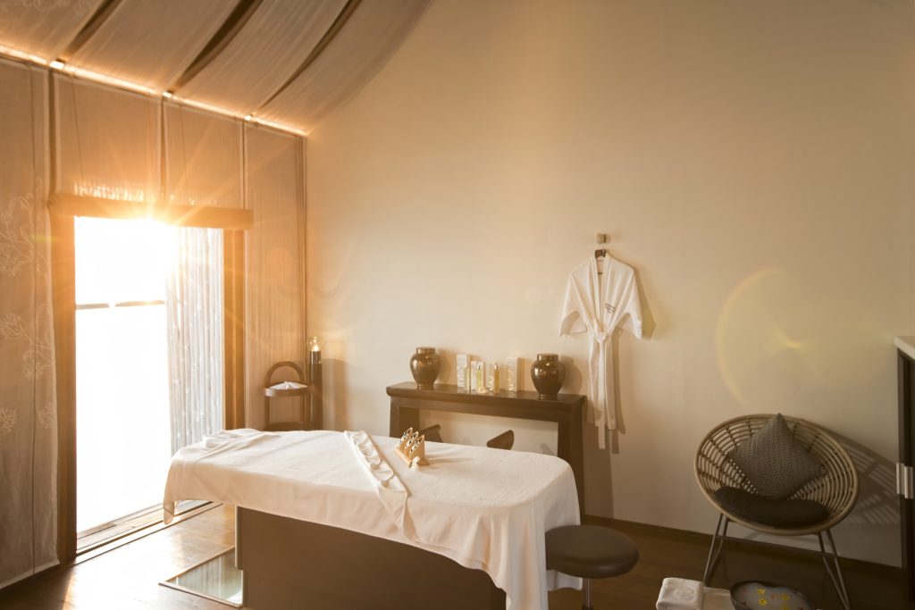 Wellnessraum im Constance Moofushi mit Massagetisch, weißem Bademantel, Holztisch und Tageslichtfenster