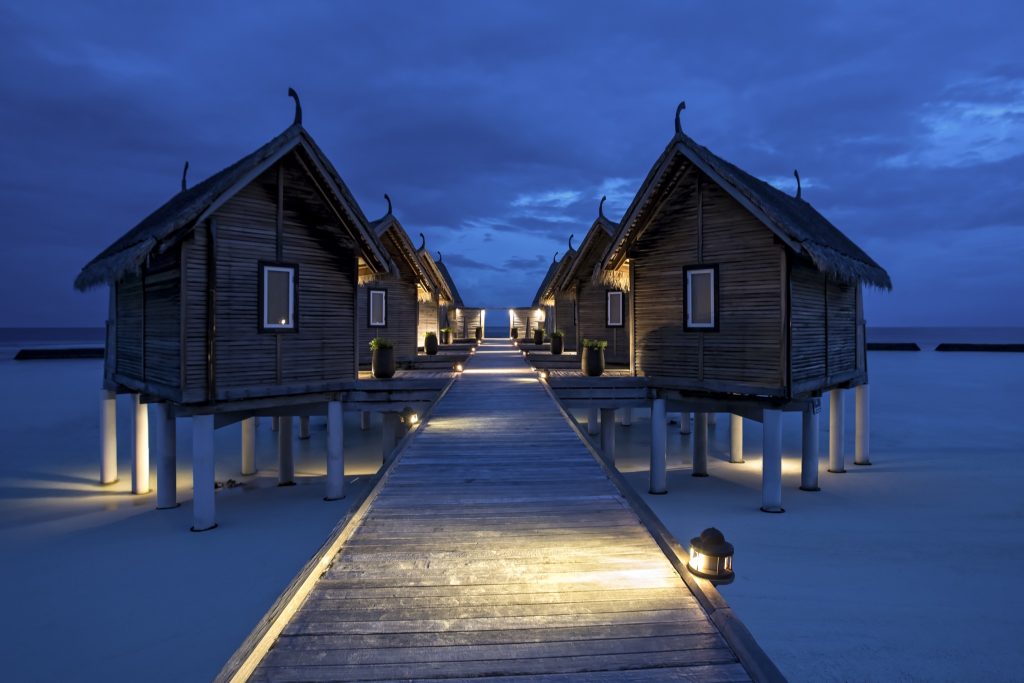 Holzbungalows auf Stelzen über dem Meer mit traditionell geformten Dächern und beleuchtetem Steg bei Dämmerung
