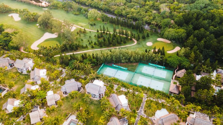 Luftaufnahme des Dinarobin Beachcomber Golf Resort & Spa mit Tennisplätzen, Strohdachgebäuden und Golfplatz in Mauritius