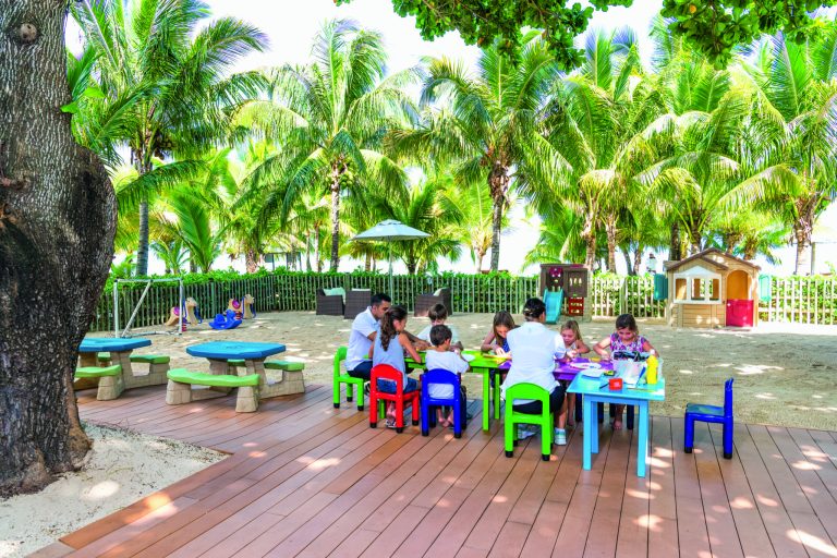 Außenbereich mit Holzdielen, bunten Tischen und Stühlen, spielende Kinder und Palmen im Hintergrund in Mauritius