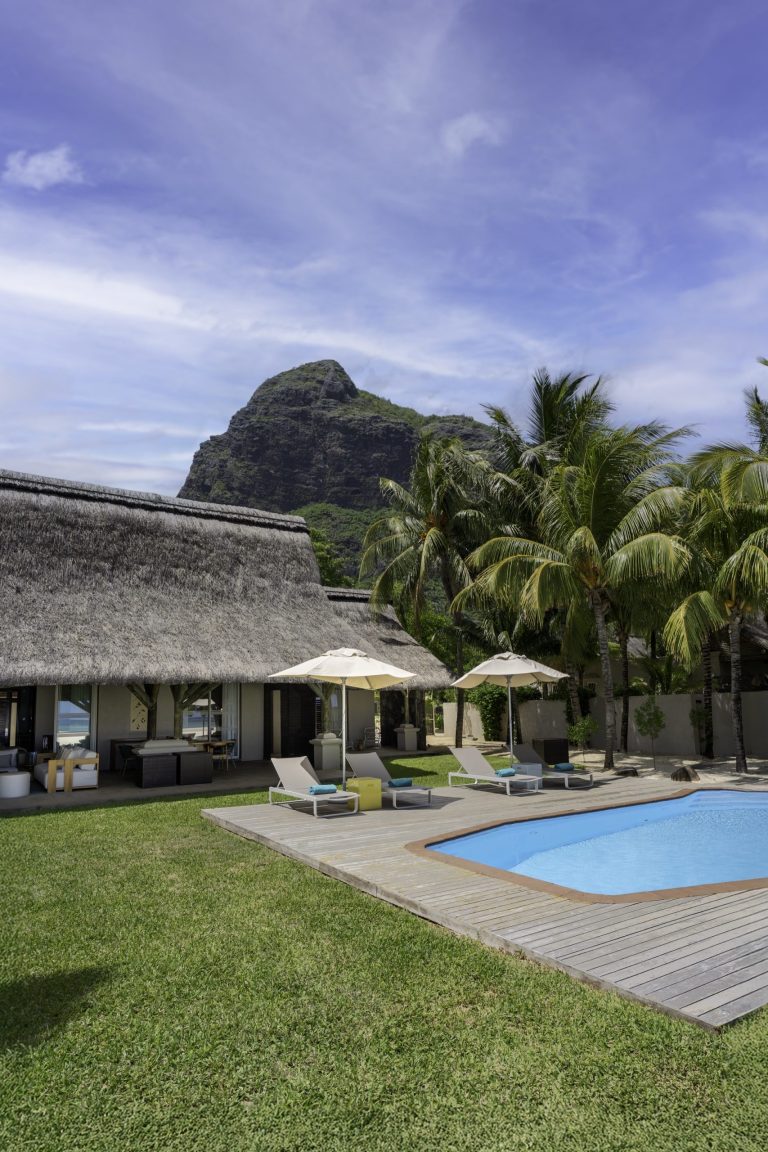 Außenbereich mit rechteckigem Pool, hölzernem Deck, Liegestühlen unter weißen Sonnenschirmen und Bergkulisse auf Mauritius