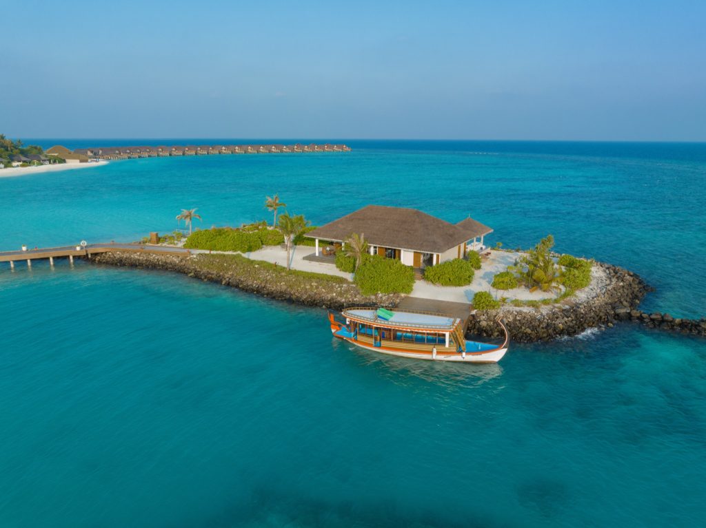 Resort auf kleiner Insel mit Holzsteg, traditionellem Boot und zentralem Gebäude mit Naturdach, Malediven