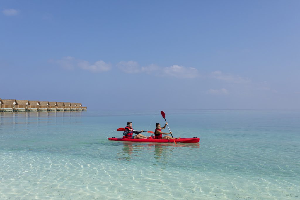 Zwei Personen paddeln in einem roten Kajak auf flachem, klarem Wasser vor Stelzen-Holzhütten am Emerald Faarufushi Resort &.
