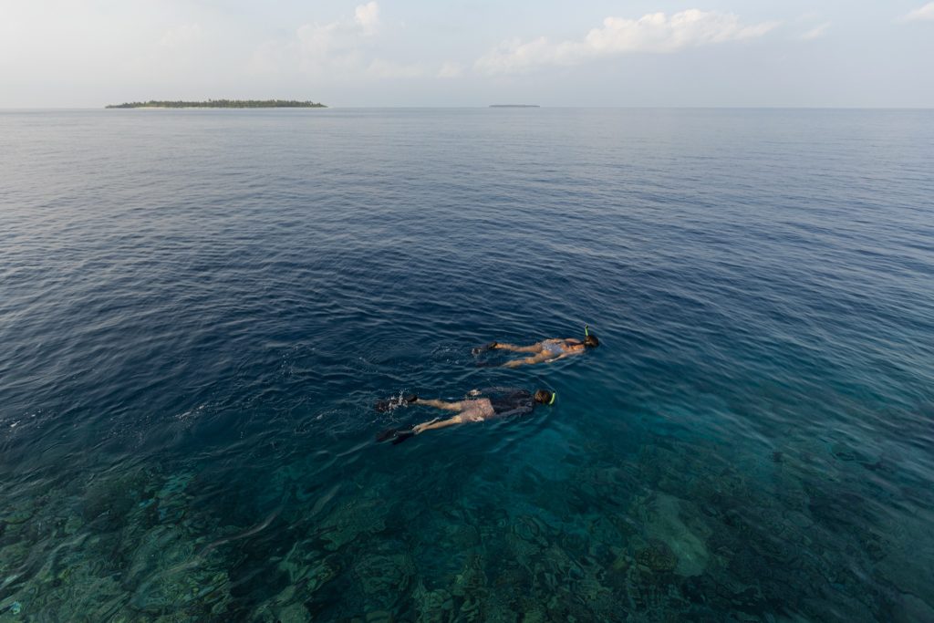 Zwei Personen schnorcheln im klaren blau-grün schimmernden Wasser vor einer schmalen Insel am Horizont