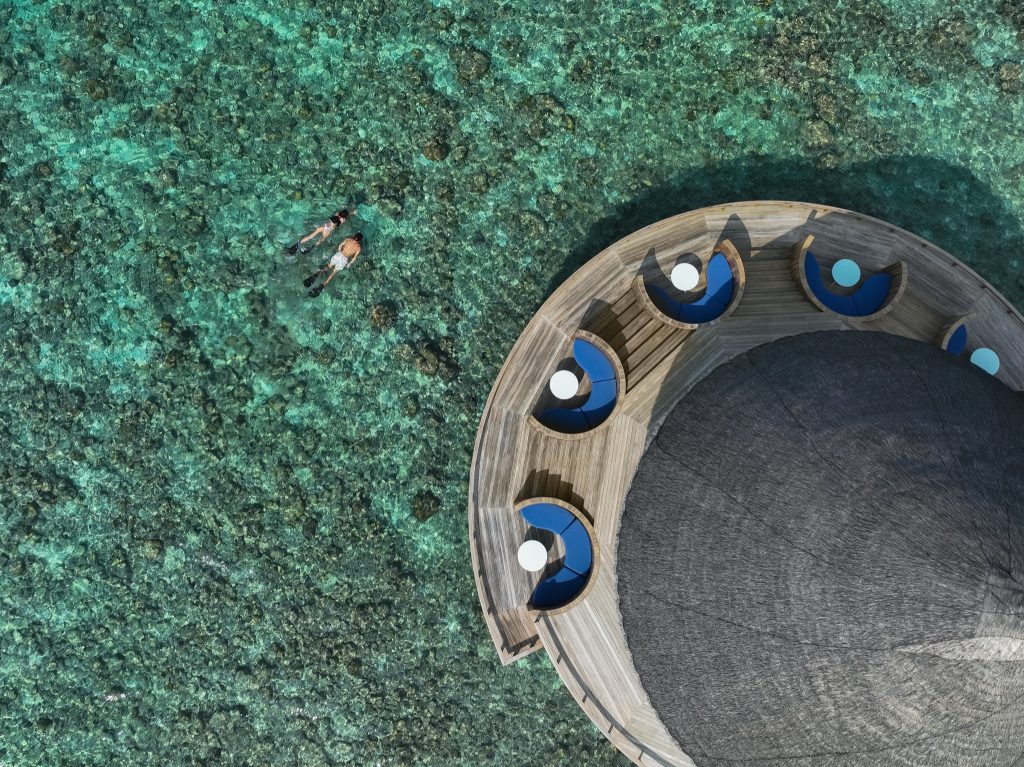 Vogelperspektive auf rundes Holzdeck mit blauen Sitzkissen und weißen Tischen über klarem türkisfarbenem Wasser