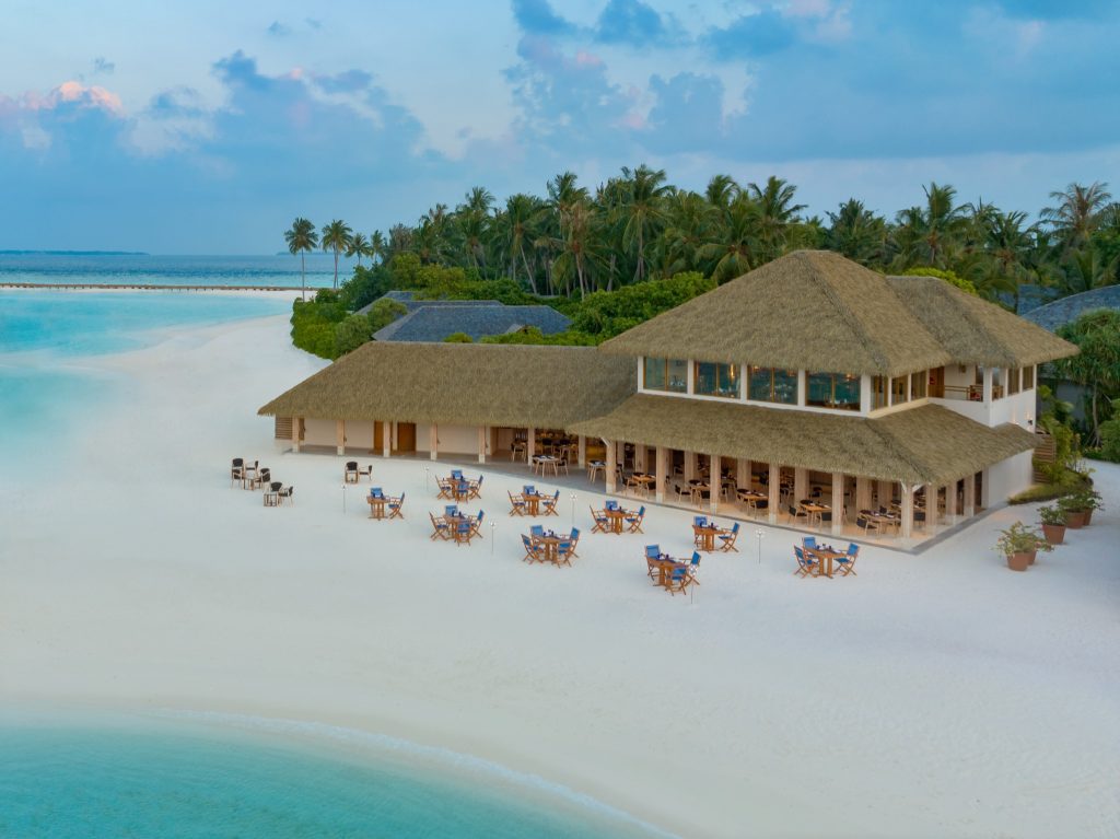 Strandhotel mit Reetdach, Holzstühle mit blauen Polstern auf weißem Sand und tropischer Vegetation im Hintergrund