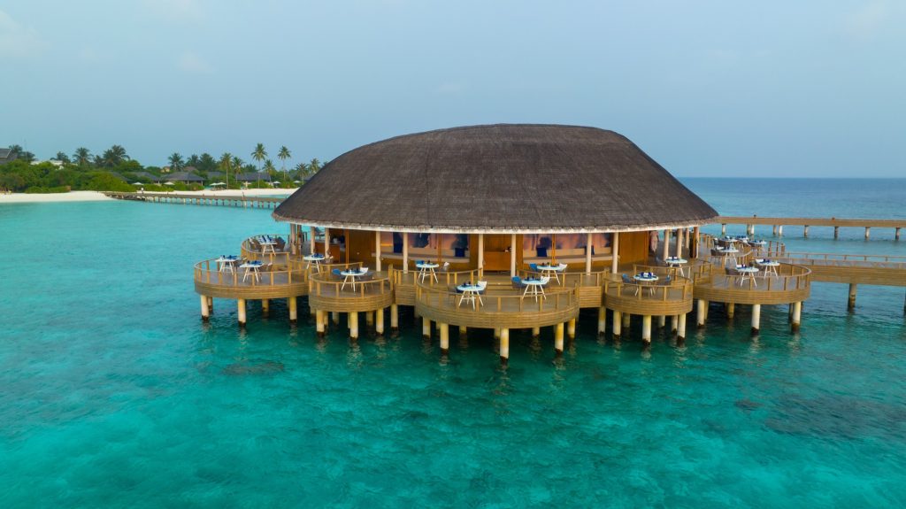 Rundgebäude mit Palmwedeldach und Terrasse mit Tischen und Stühlen über türkisfarbenem Wasser