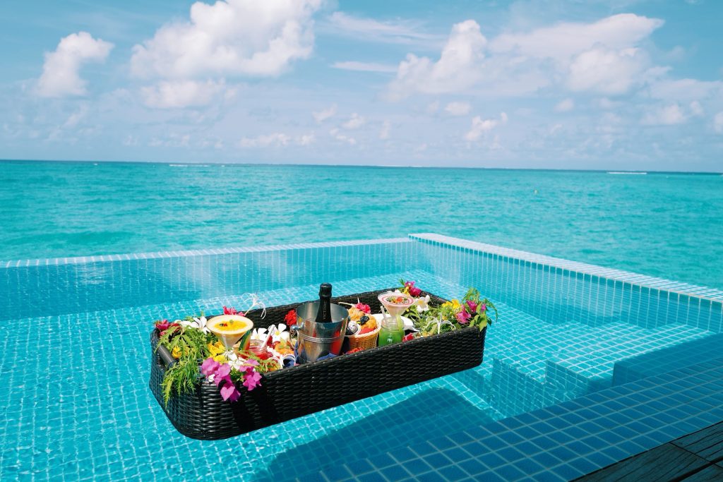 Infinity-Pool mit schwimmendem Tablett aus Korbmaterial, Speisen, Blumen, Champagner und Meer im Hintergrund