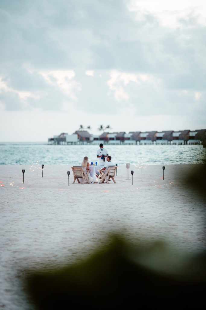 Gedeckter Holztisch mit Stühlen am Strand, Meer und Pfahlbauten im Hintergrund, Fackeln beleuchten den Bereich