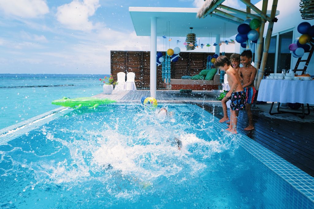Drei Kinder am Beckenrand eines Pools mit hölzernem Terrassendeck und offenem Meer im Hintergrund