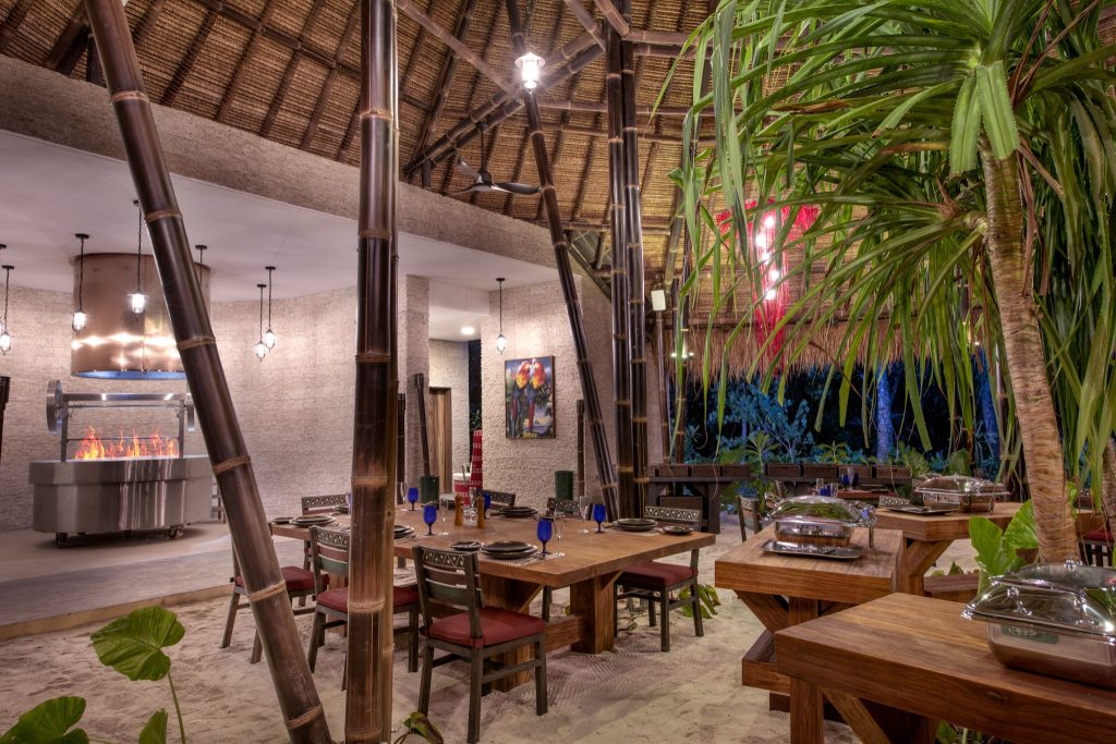 Restaurant Amazonico mit Schilfdach, Holztischen und roten Kissen auf Sandboden im Emerald Maldives Resort & Spa