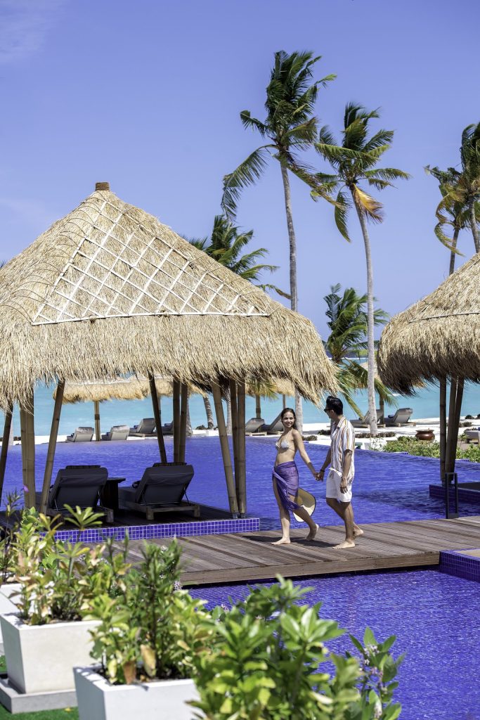 Poolbereich mit strohbedeckten Pavillons, Holzliegen und Palmen im Emerald Maldives Resort & Spa