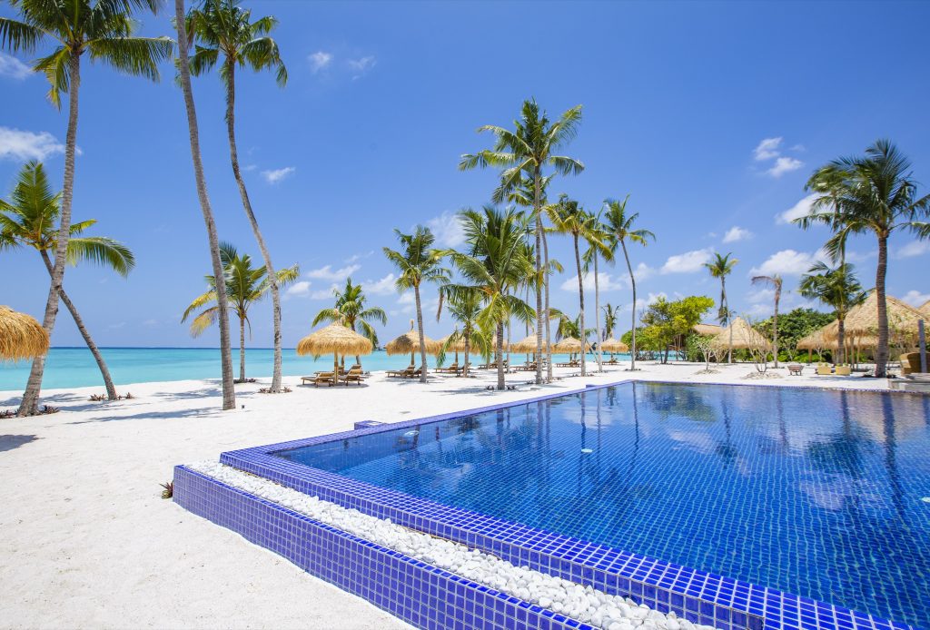 Infinity-Pool mit blauen Kacheln am weißen Sandstrand mit Palmen und Strohdachhütten auf den