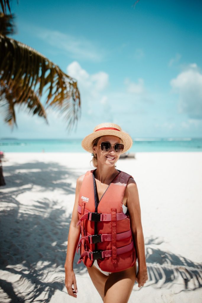 Frau mit roter Schwimmweste und Strohhut am weißen Sandstrand mit Palme und türkisblauem Meer im Emerald Maldives Resort &.