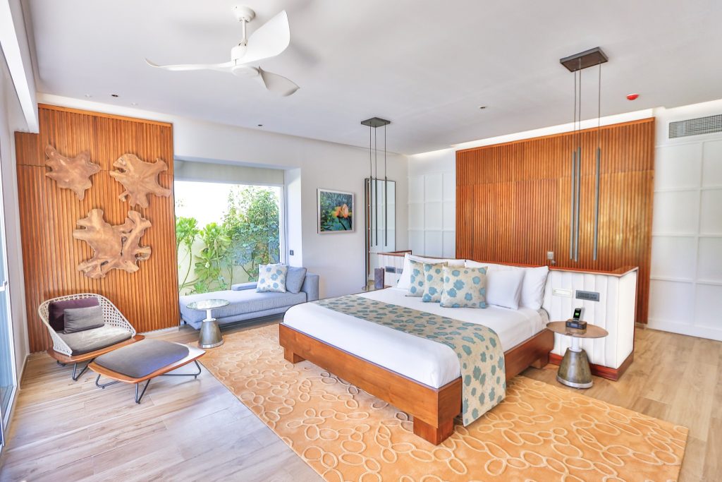 Zimmer der Beach Villa im Emerald Maldives Resort & Spa mit großem Bett, Holzwand, Sofa und Fenster mit Gartenblick