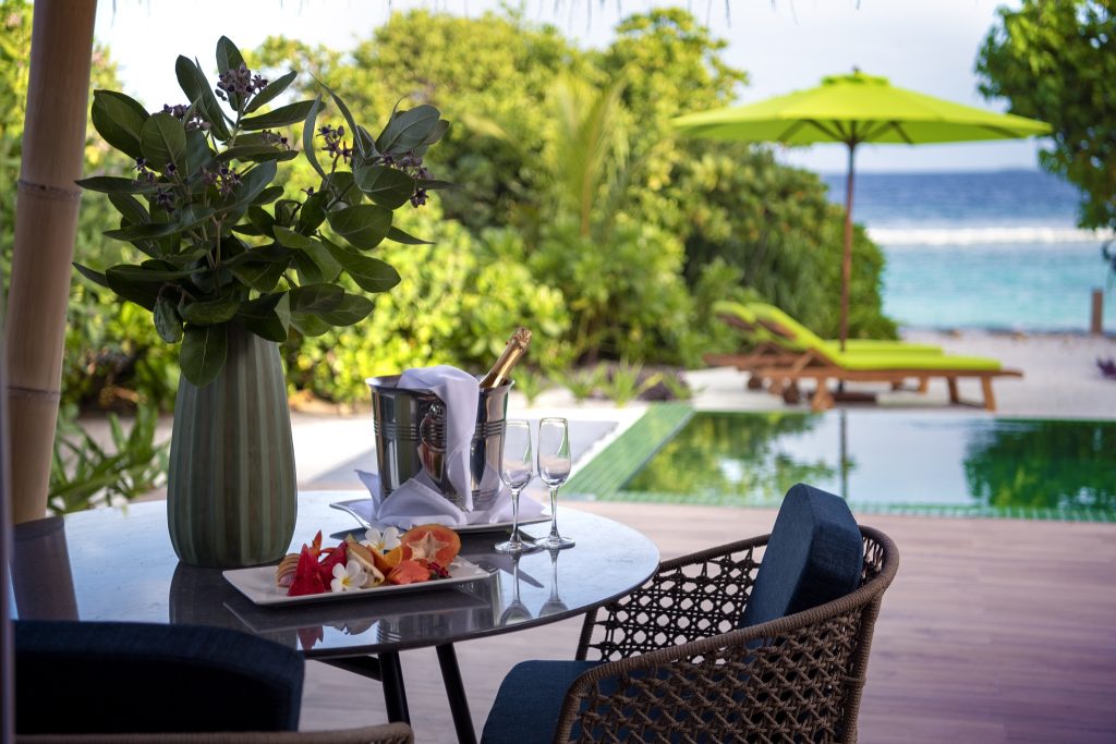 Tisch mit Vase, Sektkühler, Gläsern und Obstplatte vor Pool und grüner Landschaft im Emerald Maldives Resort & Spa
