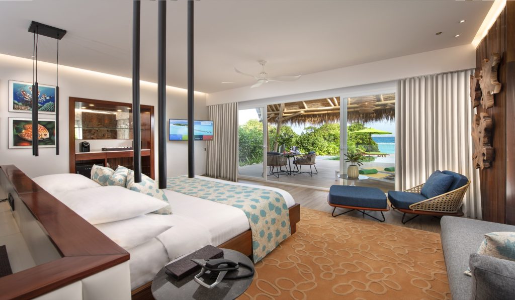 Hell eingerichtetes Zimmer in der Beach Villa mit großem Bett, Rattan-Sessel, Holzrückwand und Meerblick