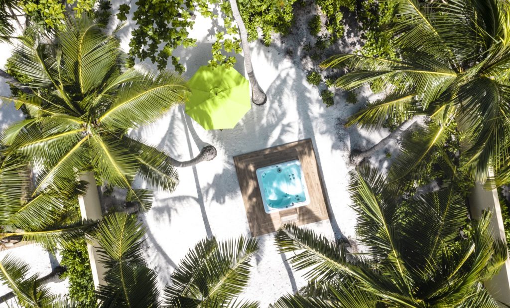 Draufsicht auf quadratischen Whirlpool auf Holzdeck mit weißem Sand, Palmen und grünem Sonnenschirm im Emerald Maldives.