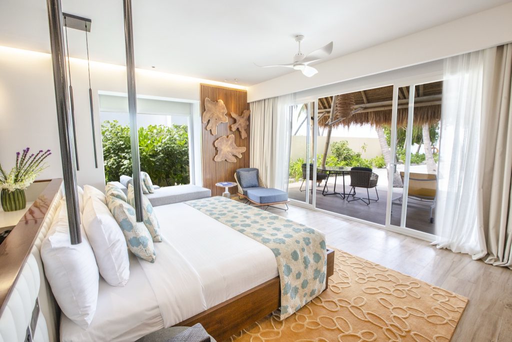Modernes Hotelzimmer Emerald Maldives Resort & Spa mit großem Bett, Holzpaneelen und Terrasse mit Blick ins Grüne