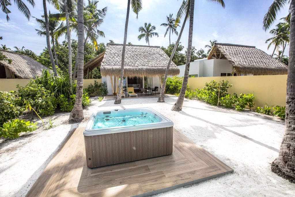 Jacuzzi auf Holzplattform vor Bungalow mit Strohdach, umgeben von weißem Sand und Palmen im Emerald Maldives Resort & Spa
