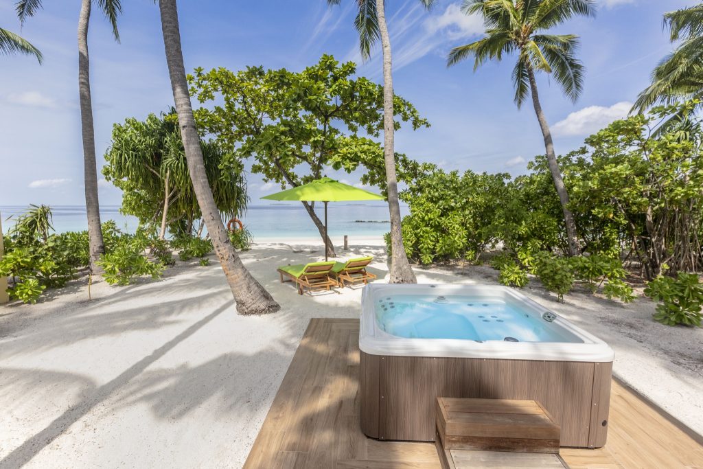 Jacuzzi auf Holzplattform am Strand mit grünen Liegestühlen und Sonnenschirm, Palmen und Meer im Hintergrund