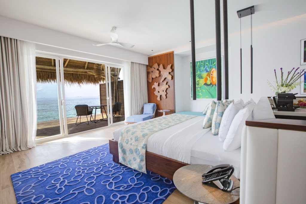 Modernes Hotelzimmer der Wasser-Villa mit großem Bett, blauem Teppich, Balkon und Meerblick im Emerald Maldives Resort & Spa