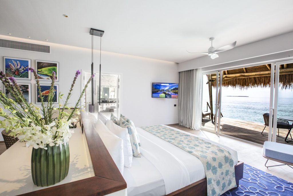 Hotelzimmer mit großem Bett, weißer Bettwäsche, türkis gemustertem Überwurf, Holzterrasse und Meerblick im Emerald Maldives.