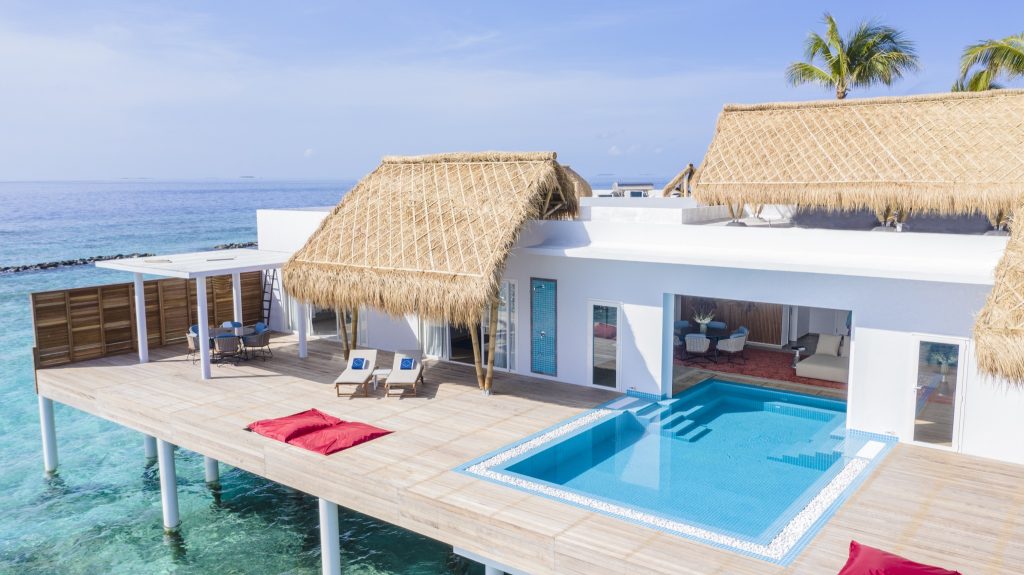 Moderne Überwasservilla auf Pfählen mit Holzterrasse, strohgedecktem Dach, Pool und Sitzgruppe über türkisfarbenem Wasser