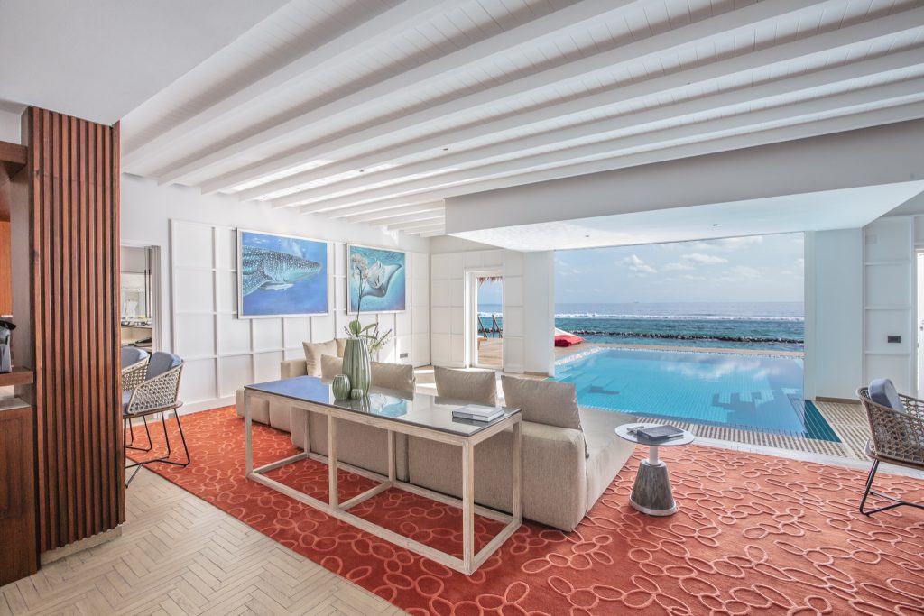 Offener Wohnbereich mit beiger Couch, Glastisch auf rotem Teppich und Meerblick in Presidential Water Villa