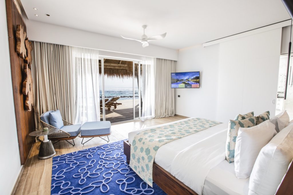Modernes Hotelzimmer mit großem Bett, blauer Sitzgruppe und bodentiefer Glasfront zur Terrasse mit Meerblick