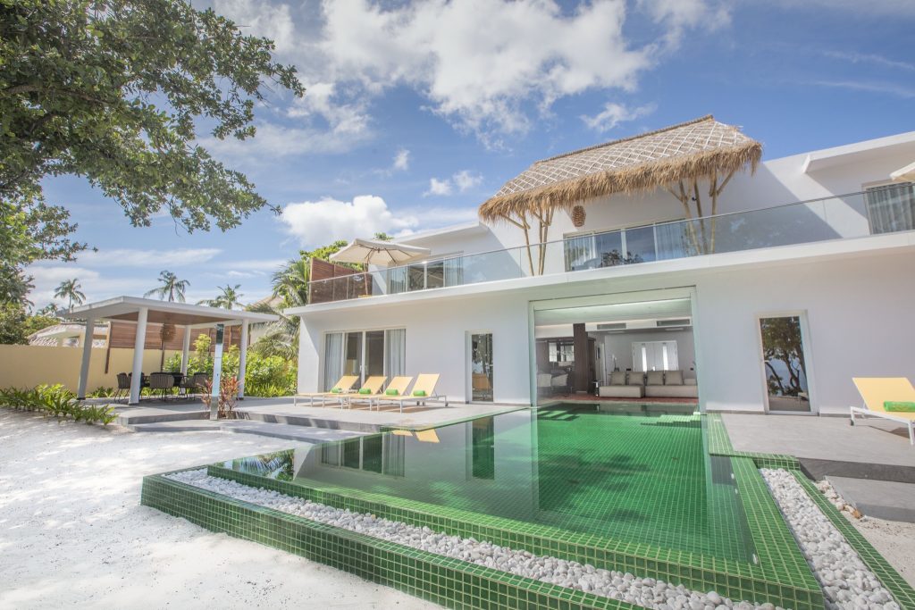 Rechteckiger Pool mit grünen Fliesen vor Royal Beach Villa, umgeben von weißen Kieselsteinen und gelben Liegestühlen