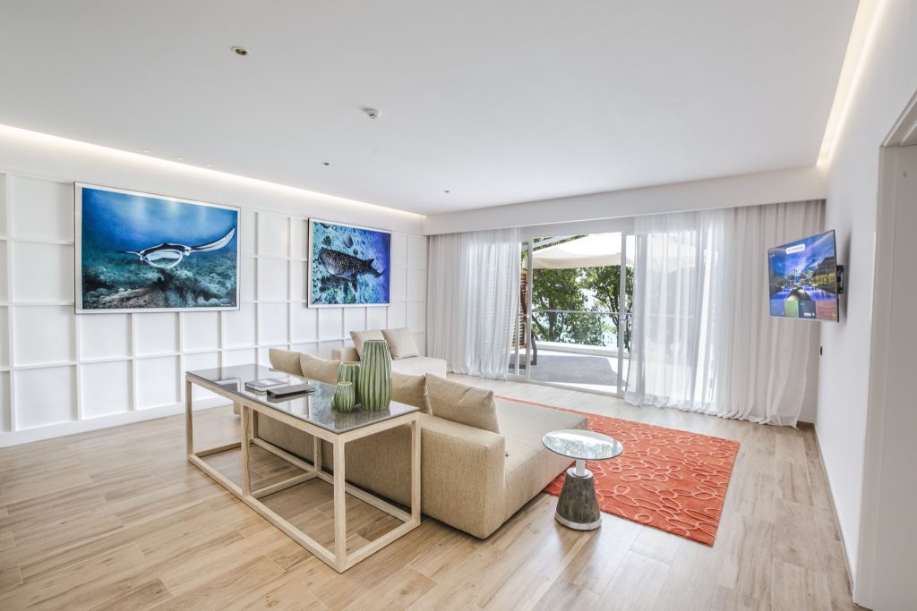 Modernes Hotelzimmer Royal Beach Villa mit hellem Holzboden, beigem Sofa, Glas-Metall-Tisch und Unterwasserbildern.