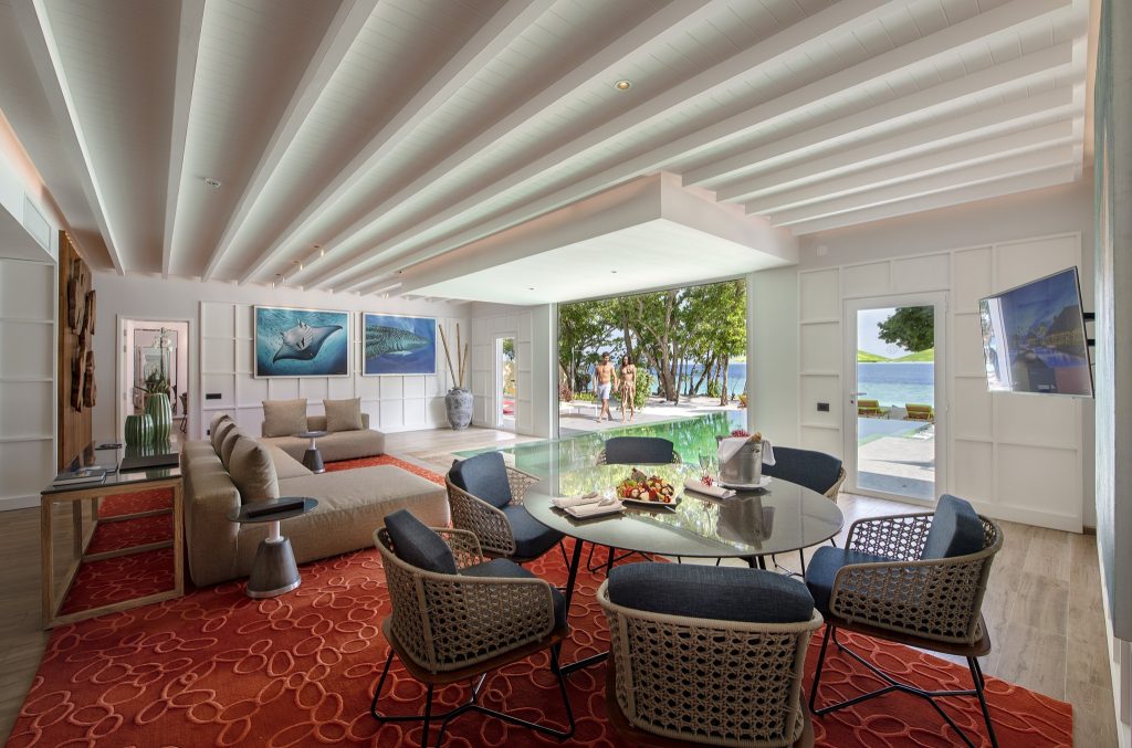 Wohnraum der Royal Beach Villa mit Glastisch, Stühlen, großem Fenster und Blick auf Pool und Meer