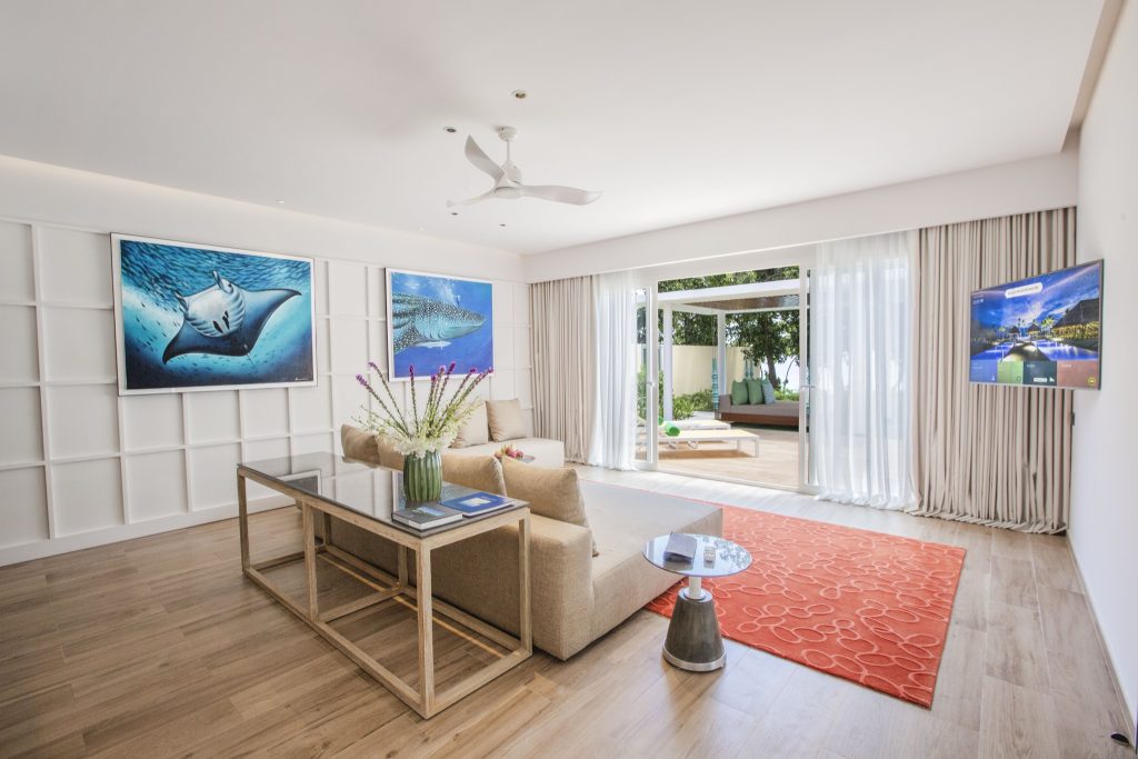 Helles Wohnzimmer der Superior Beach Villa mit beigen Möbeln, Glastisch, ozeanische Bilder und Blick auf Terrasse in den.