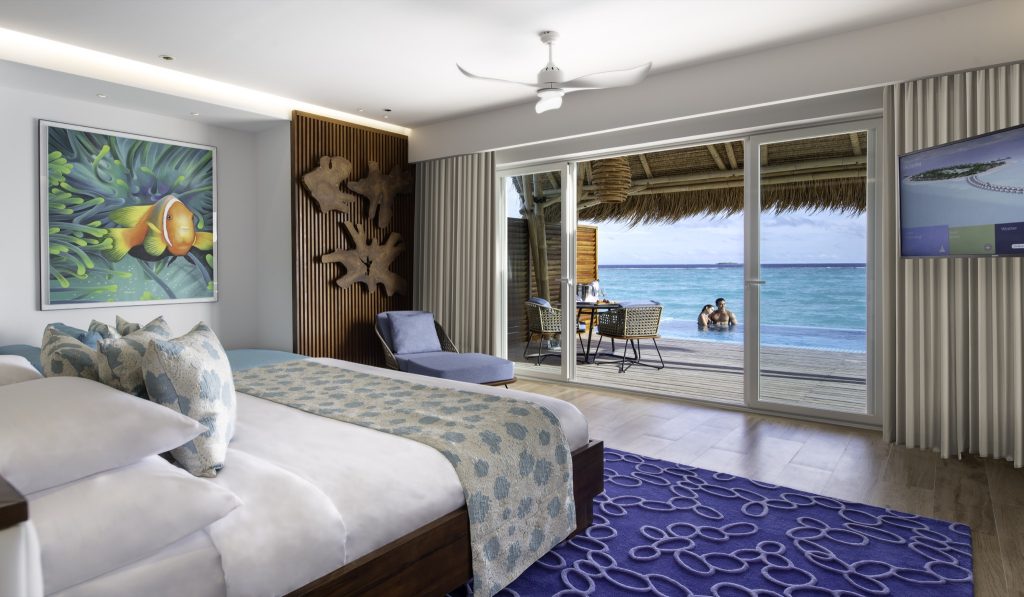 Hotelzimmer Water Villa mit großem Bett, blauem Teppich, Fischbild und Meerblick auf den Ozean