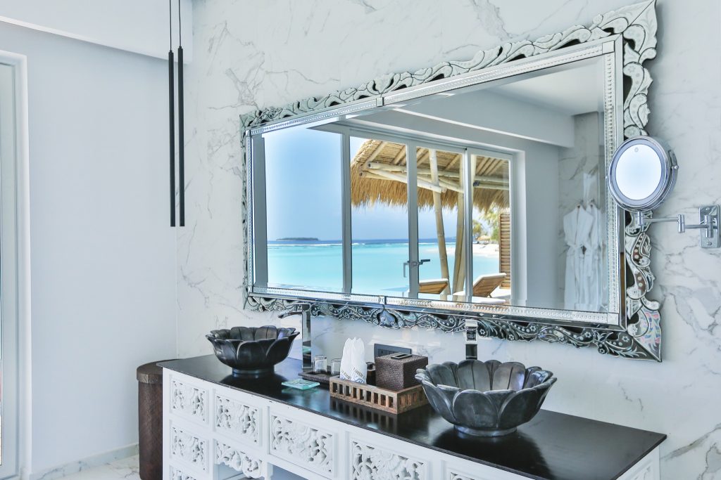 Badezimmer mit großem Spiegel, blumenförmigen Waschbecken und Blick auf türkisfarbenen Ozean im Emerald Maldives Resort & Spa