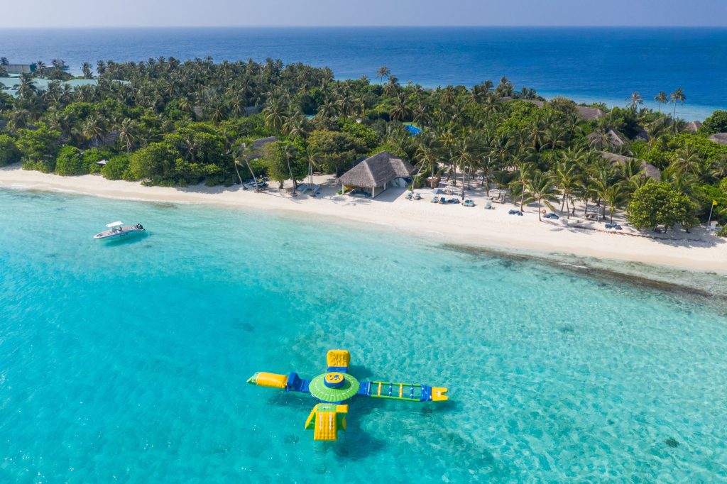 Luftaufnahme eines weißen Sandstrands mit Palmen, buntem Wasserspielgerät und Boot vor der Küste auf den