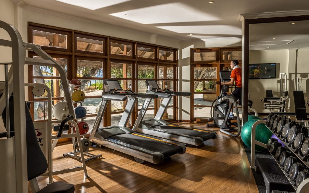 Fitnessraum mit Laufbändern, Gewichten auf Regalen, hellem Holzboden und großen Fenstern im Four Seasons Resort Maldives