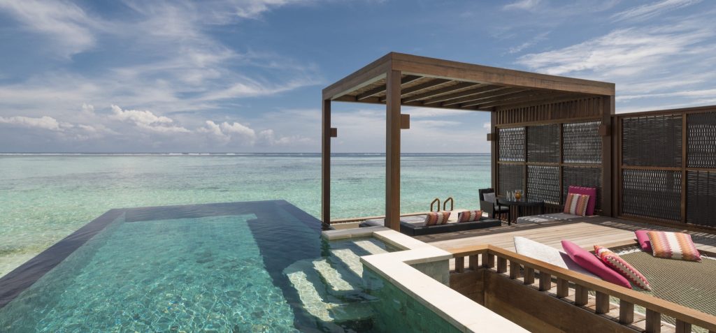 Infinity-Pool mit blauen Fliesen und hölzerne Pergola auf Terrasse mit bunten Liegekissen und Meerblick